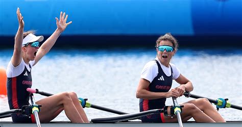 A female rower in a racing boat, focused and determined, with the Olympic rings visible in the background.